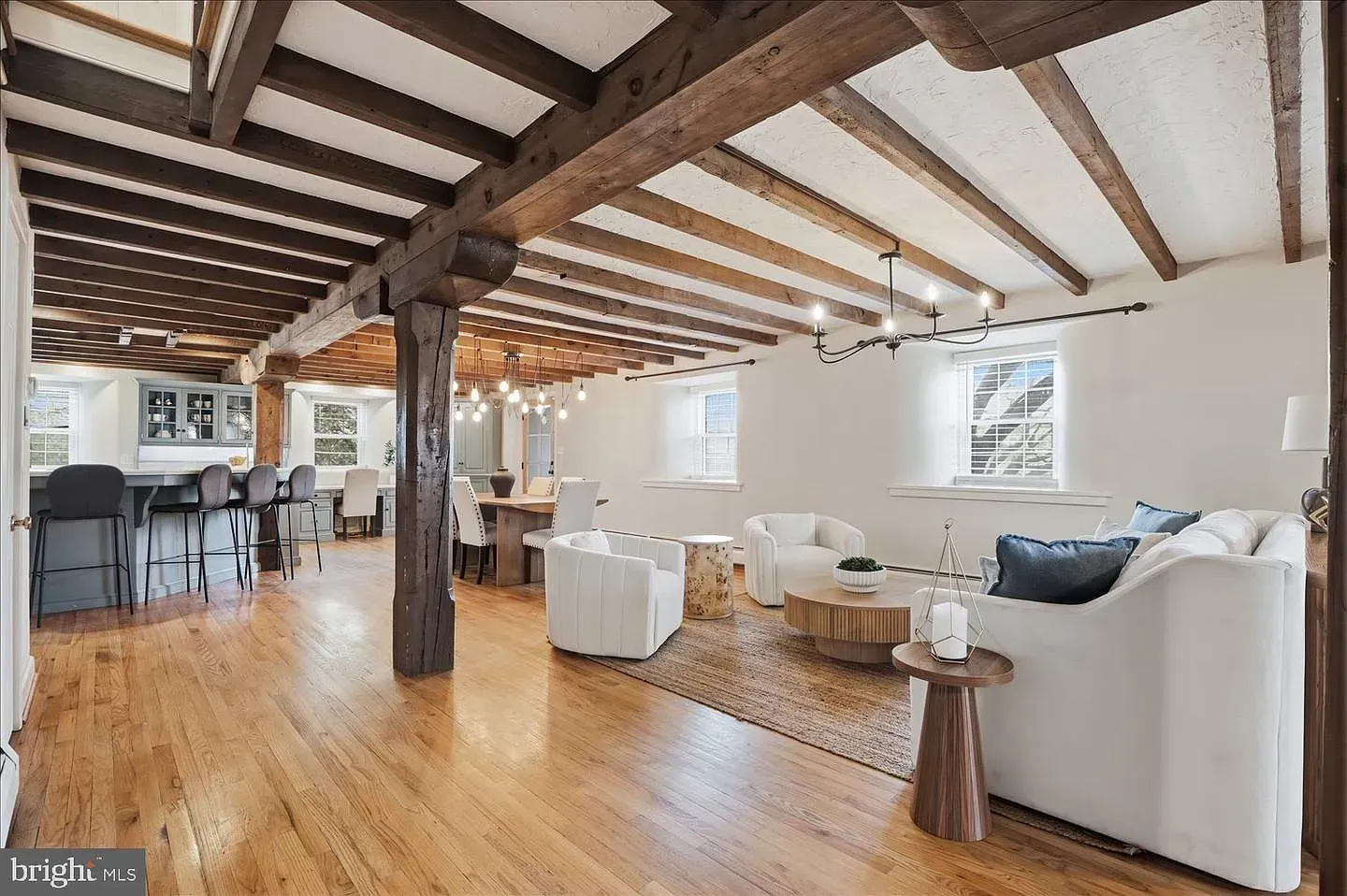 Bright open living room with exposed wood beams, hardwood floors, white sofas, and a dining area