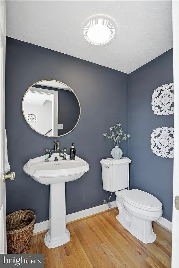 Small blue-and-white bathroom with pedestal sink, round mirror, toilet, and wood floor