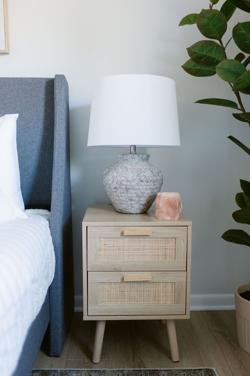 Bedroom nightstand with white lamp and woven drawers beside a bed and plant