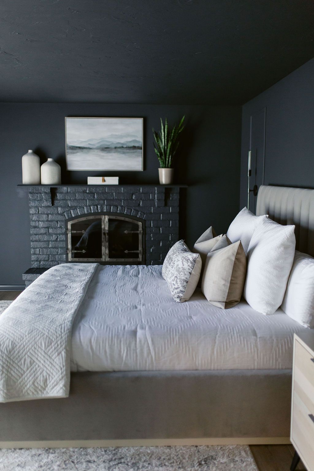 Cozy dark-gray bedroom with a neatly made bed, fireplace mantel, and framed seascape above it.