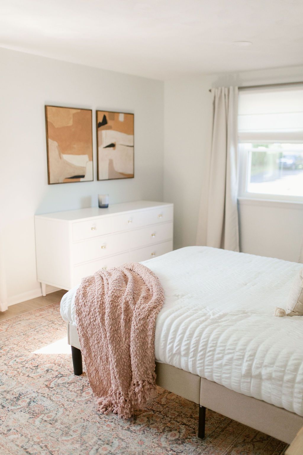 Bright bedroom with white bedding, a dresser, abstract wall art, and a pink throw blanket near a window.