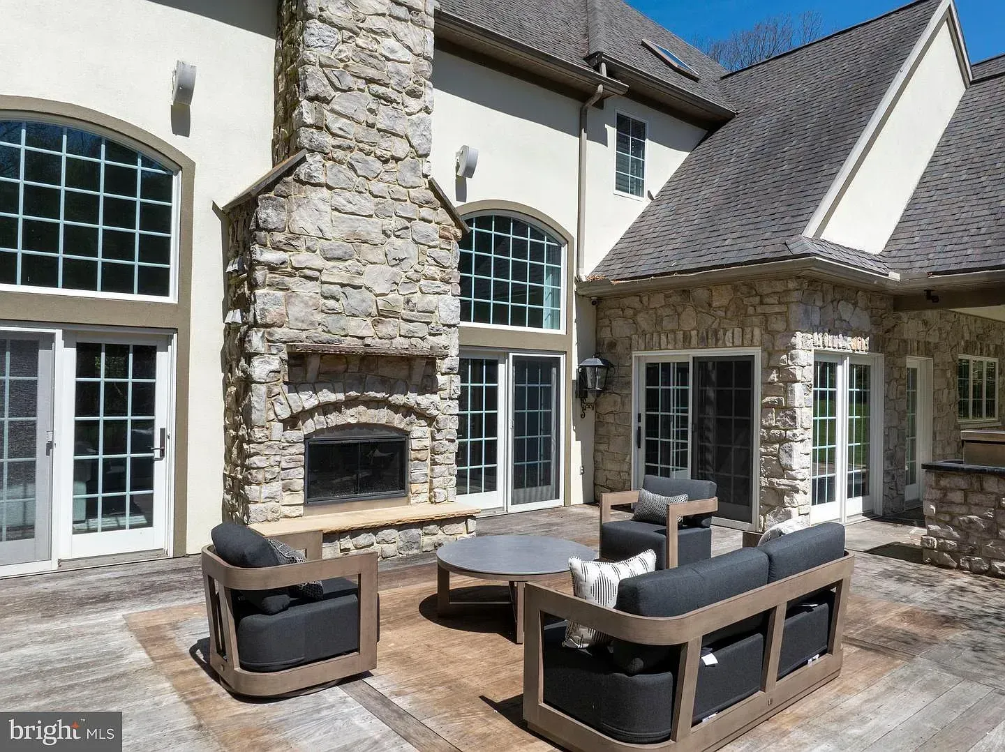 Patio with cushioned outdoor seating and a stone fireplace beside a house with large windows