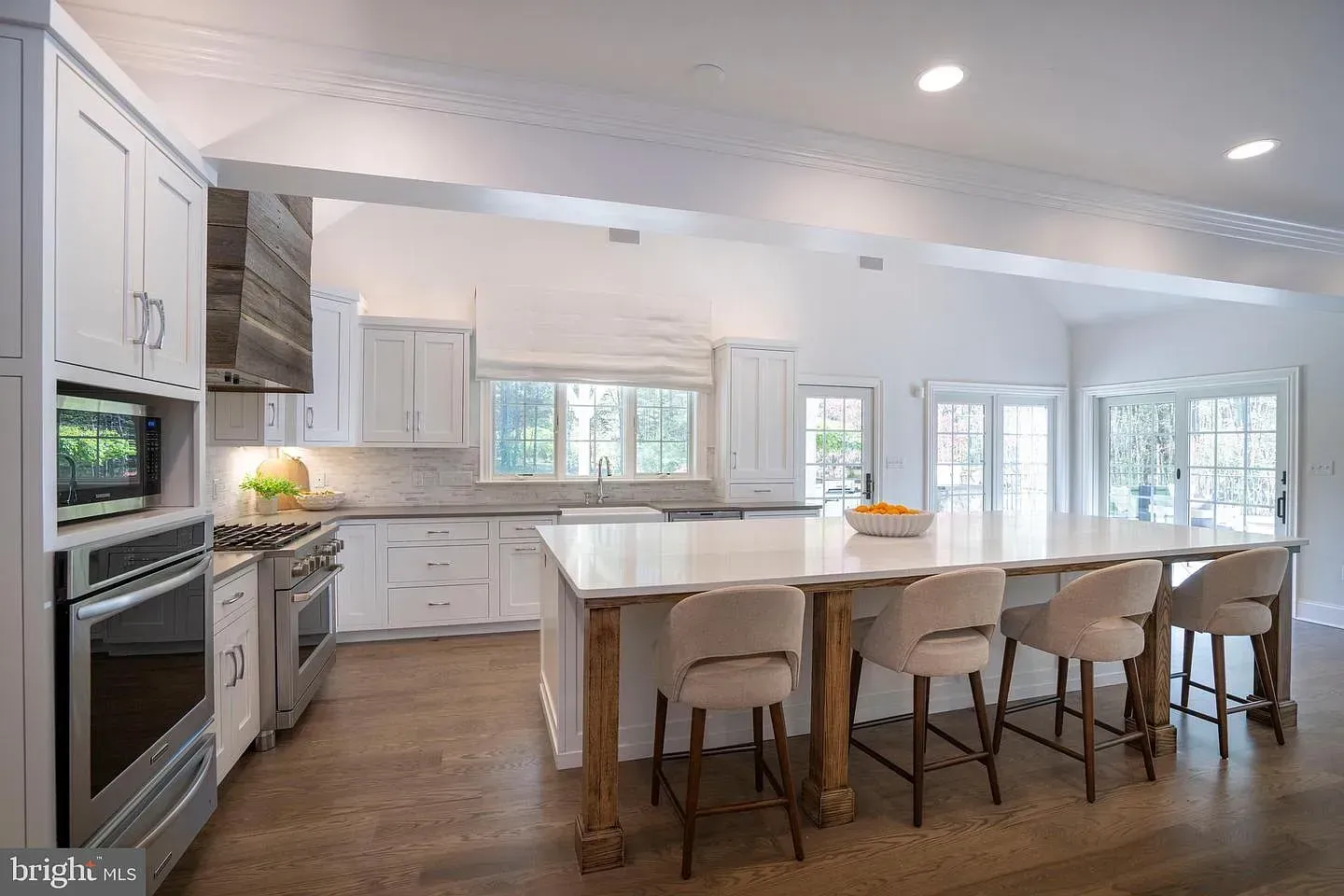 Bright white kitchen with large island, bar stools, stainless appliances, and windows overlooking trees