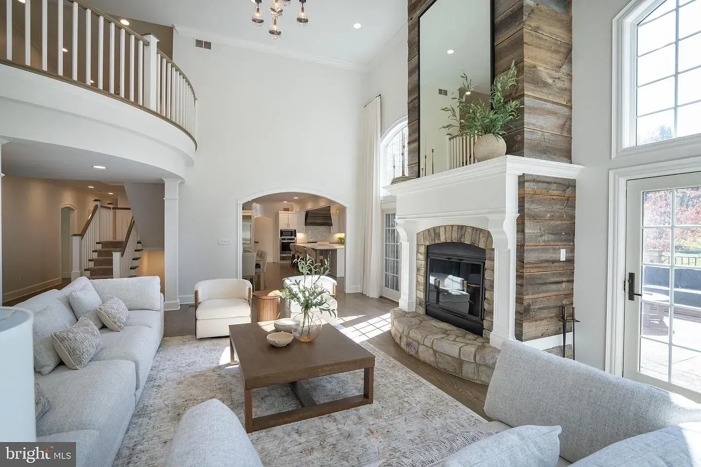 Bright, airy living room with white sofas, stone fireplace, tall windows, and a lofted balcony.