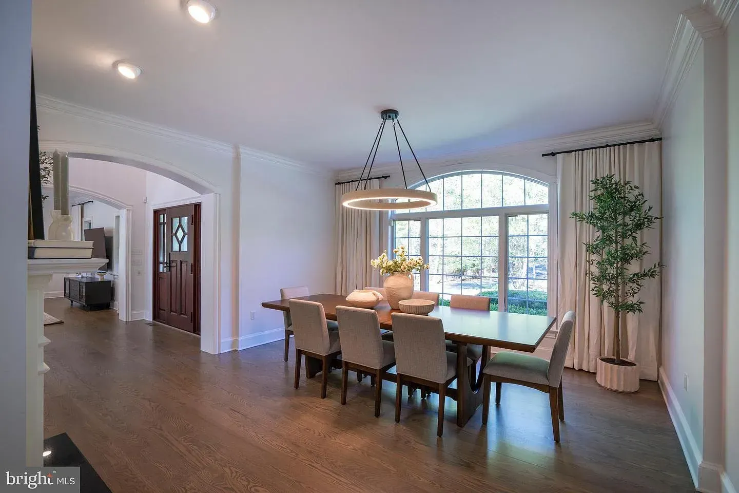 Bright dining room with a long table, beige chairs, large arched window, and potted plant.