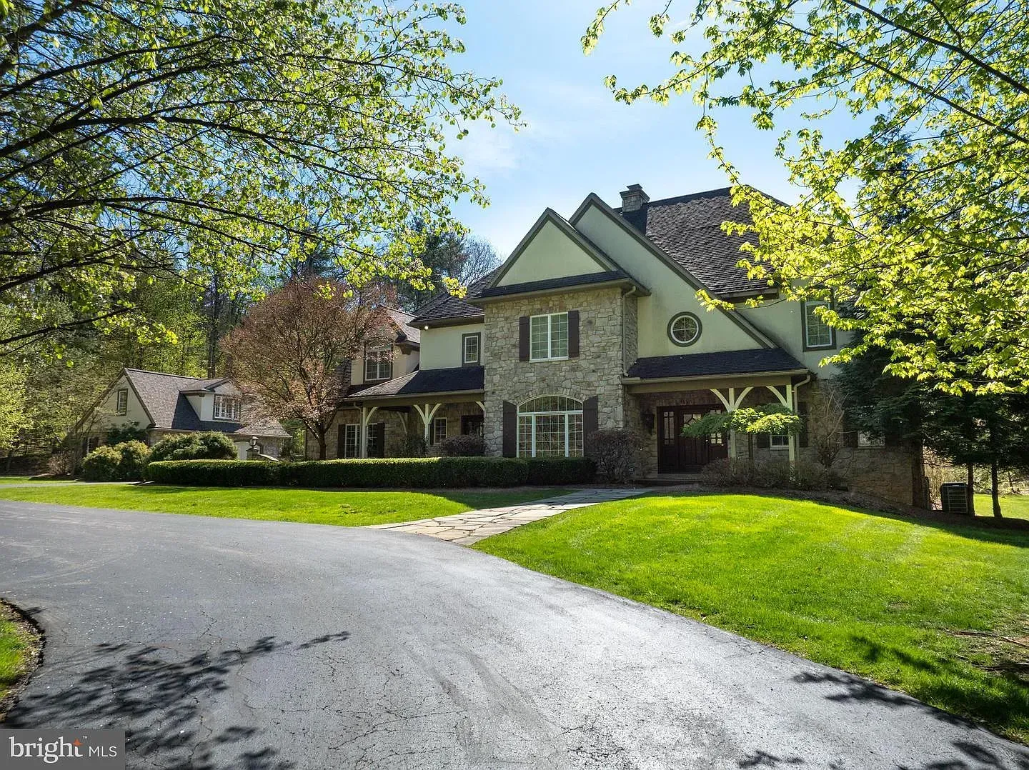 Large stone house with a driveway, green lawn, and trees in bright sunlight