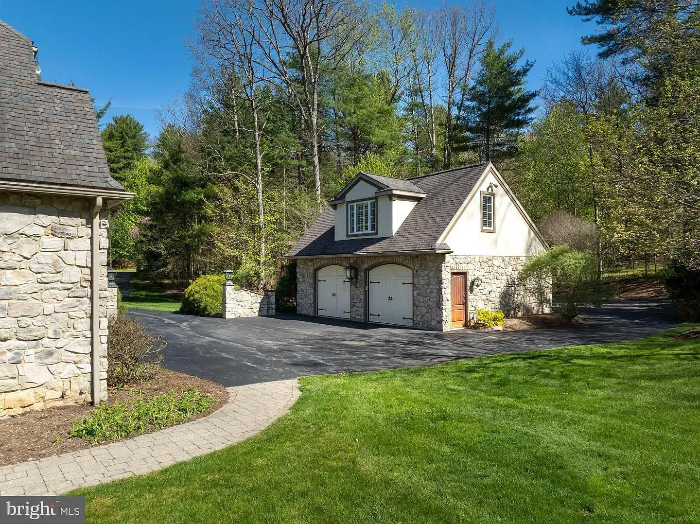 Stone garage and cottage with gravel driveway, surrounded by green lawn and trees on a sunny day