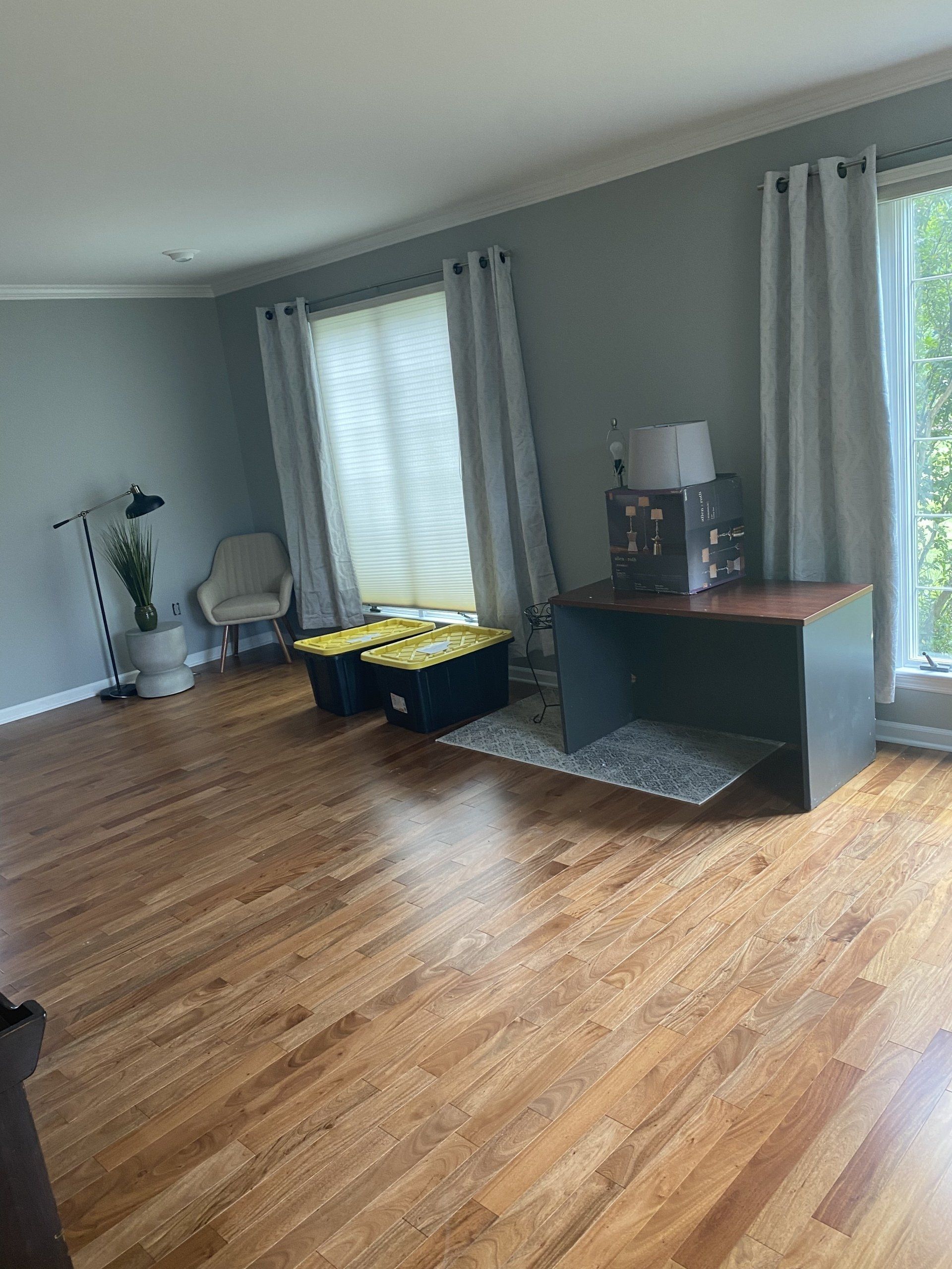 Empty living room with gray walls, wood floors, curtains, a desk, and storage bins by the window
