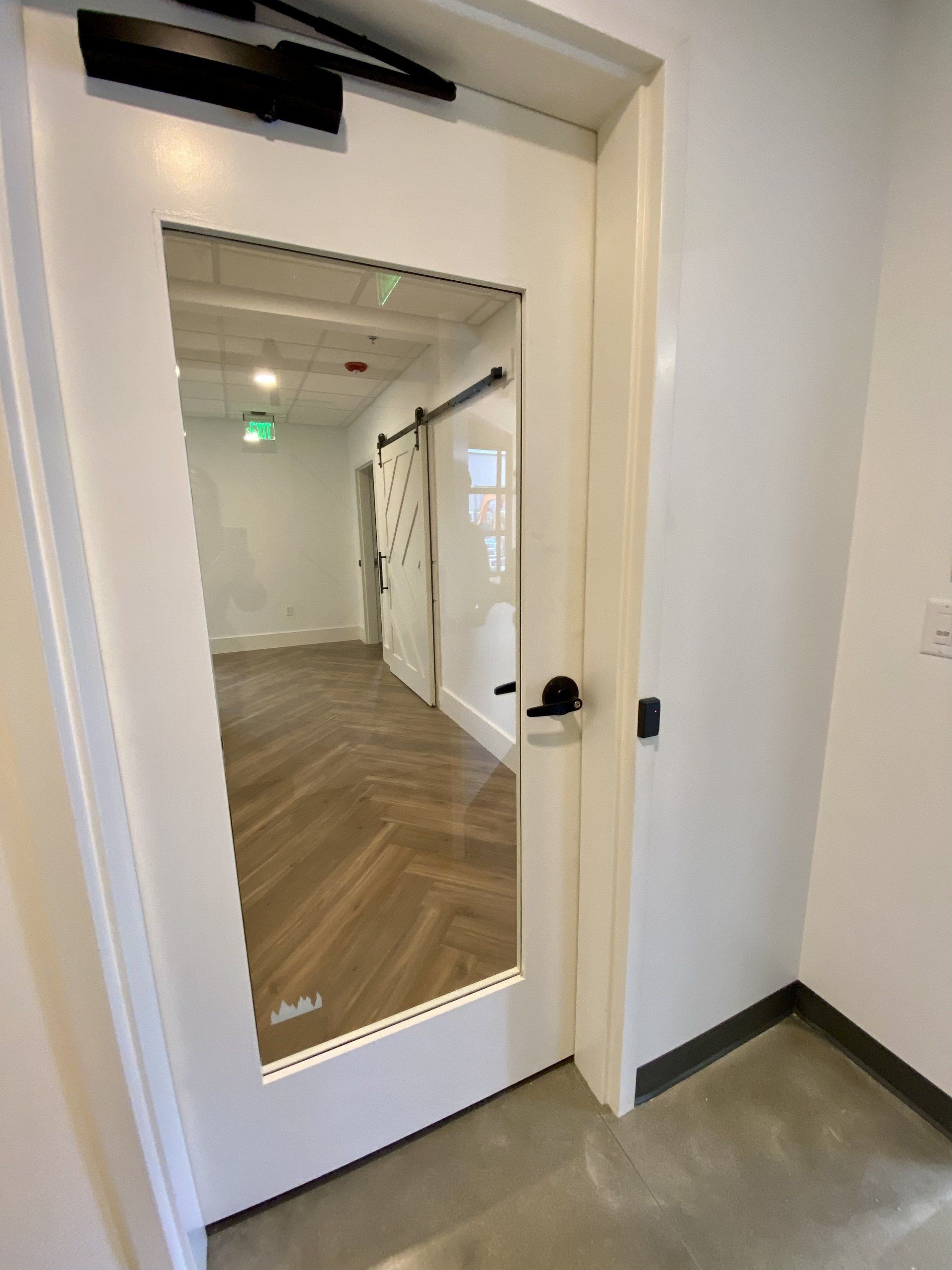 Open white door with glass panel leading to a bright office hallway with wood floors