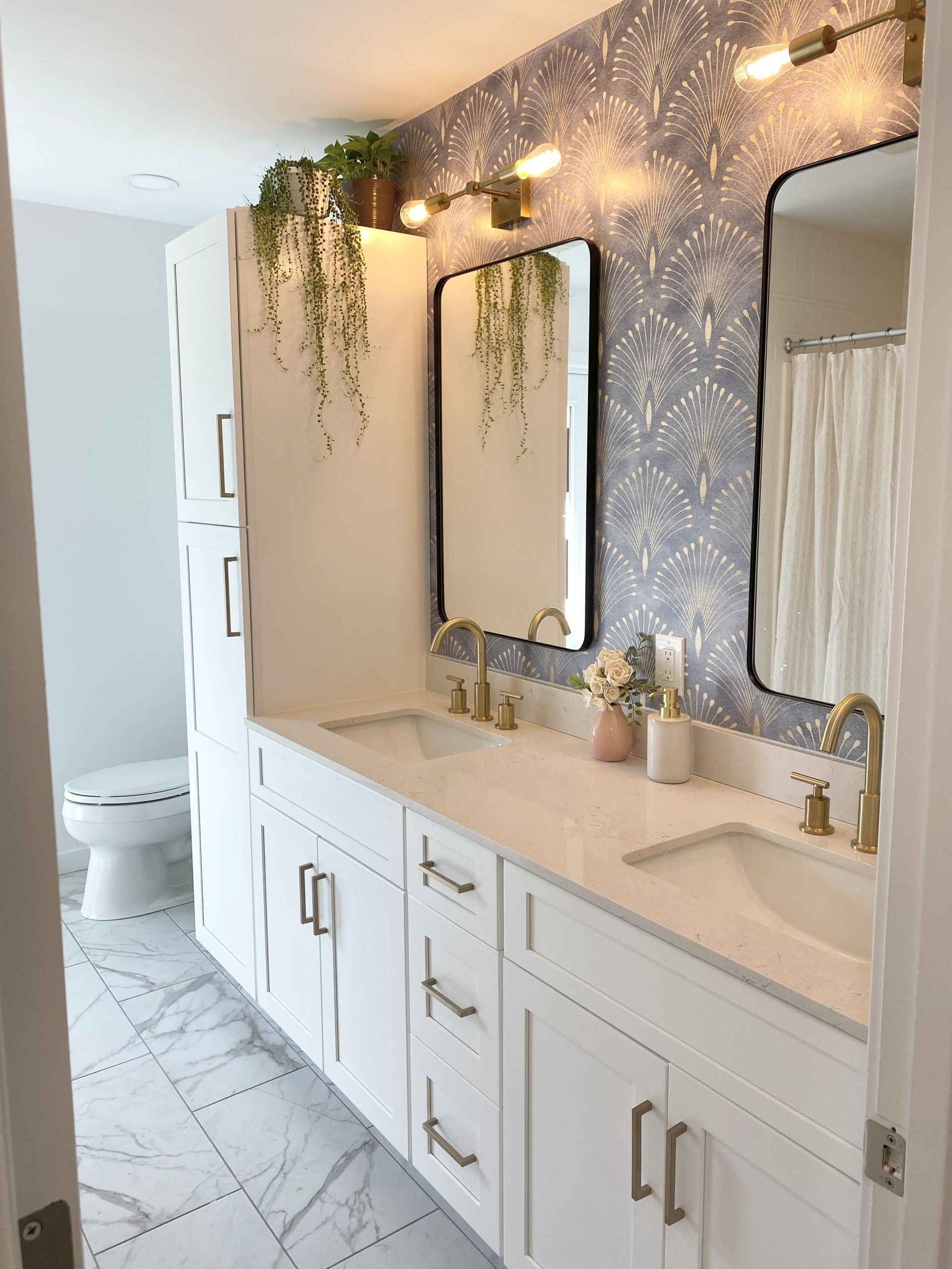 Modern bathroom with double vanity, large mirrors, wallpaper accent wall, and plants hanging above the sinks