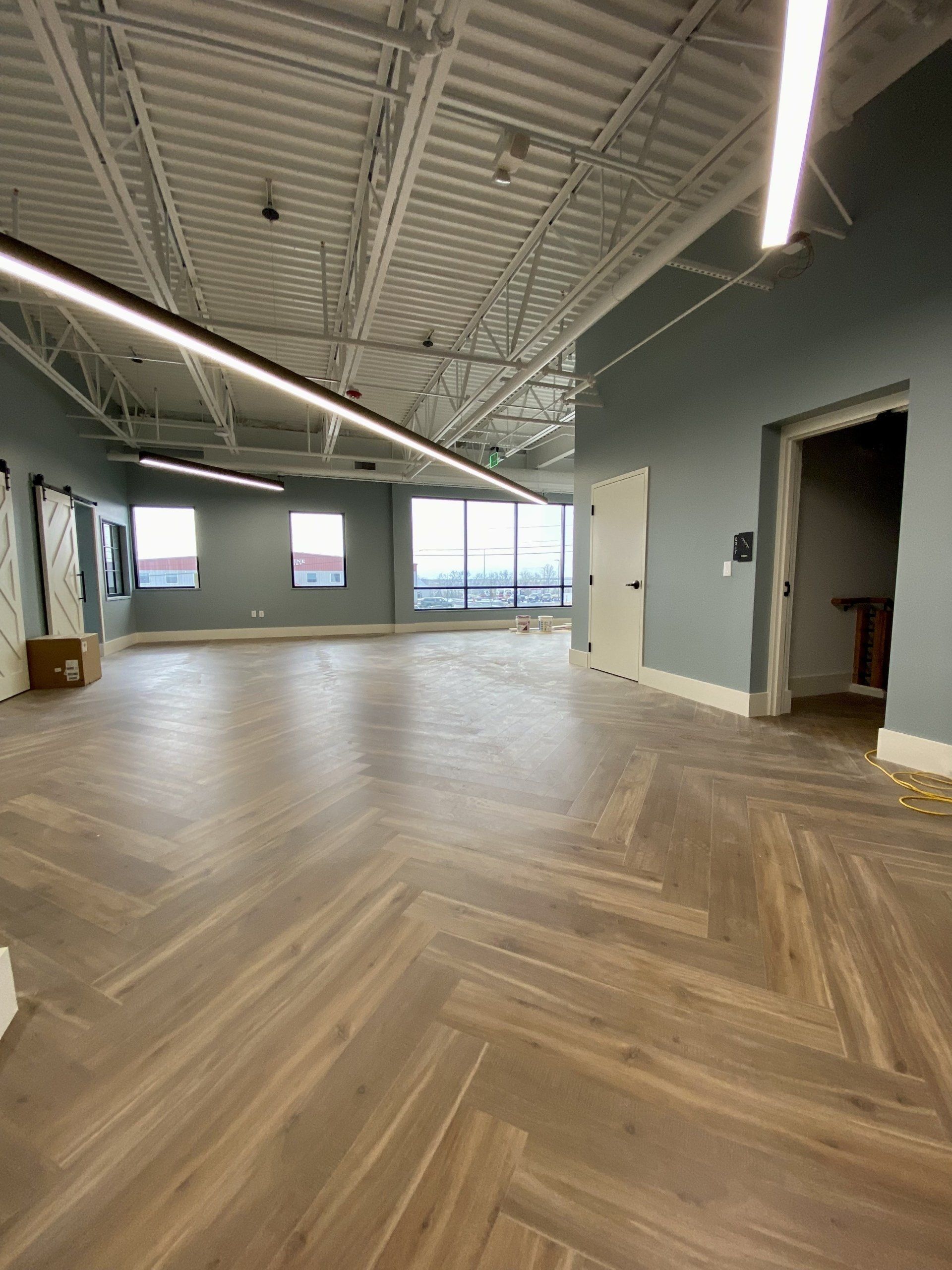 Empty modern office space with wood herringbone floor, exposed ceiling, large windows, and gray walls