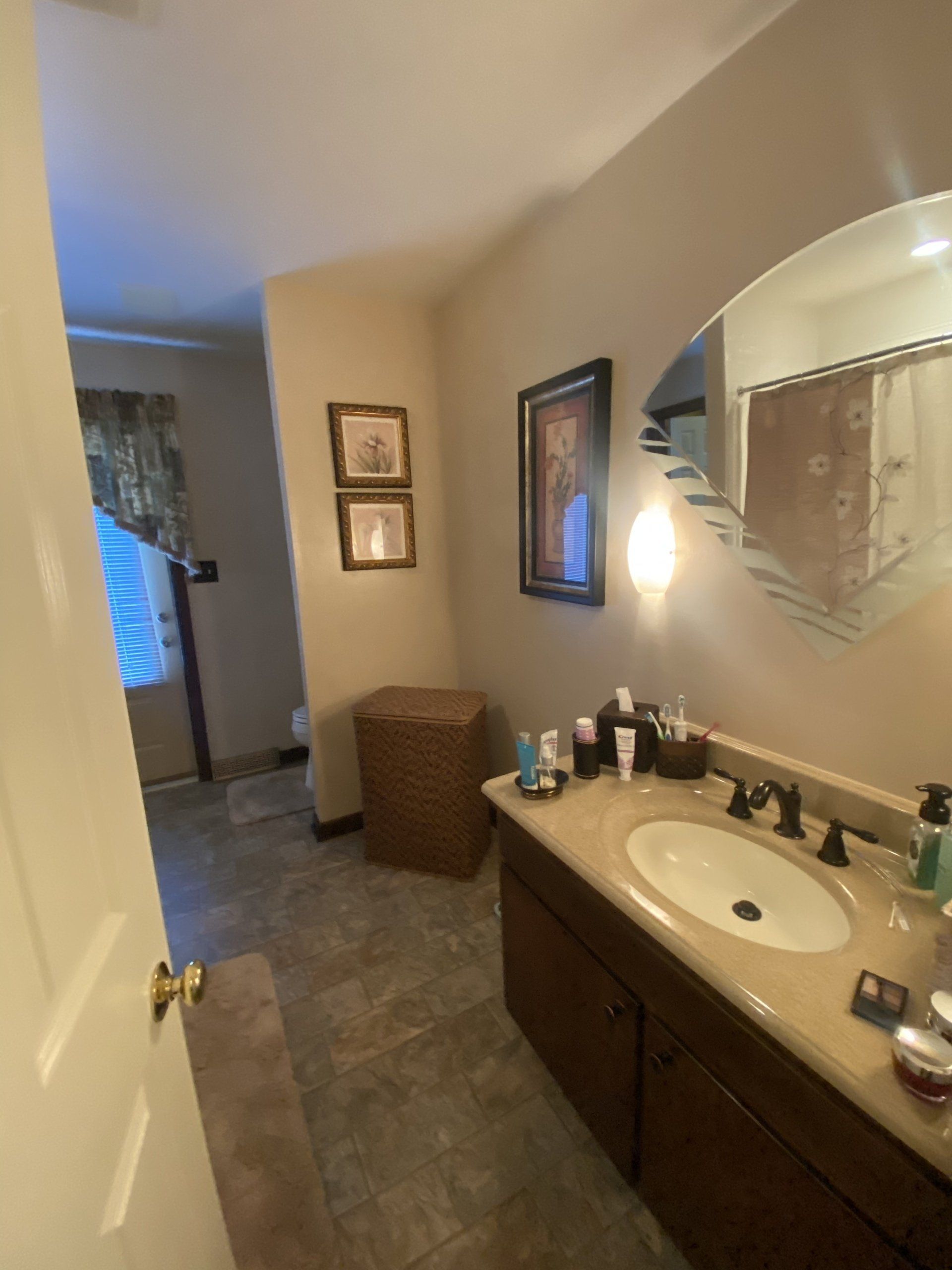 Bathroom with beige walls, wooden vanity, oval mirror, and tiled floor leading to a hallway.
