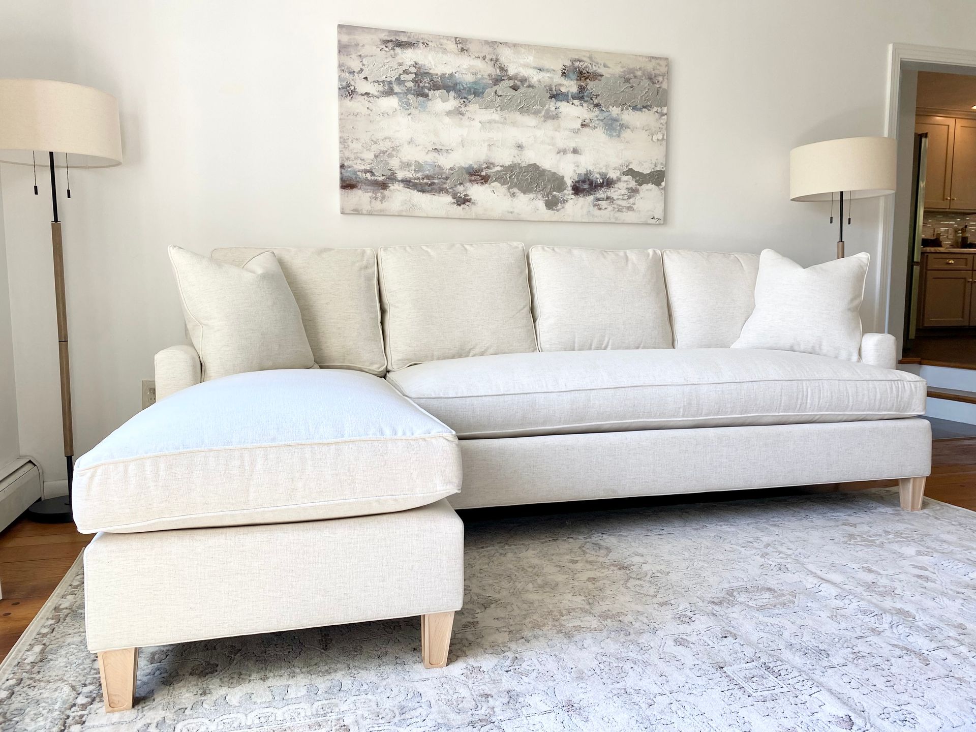 Bright living room with cream sofa, ottoman, floor lamps, and abstract wall art on a light rug