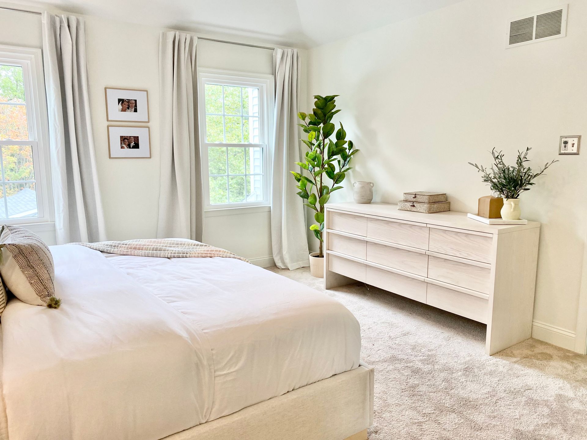 Bright bedroom with white bedding, a dresser, windows, and a potted plant on a soft carpet.
