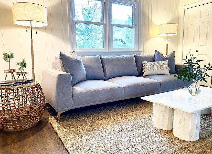 Bright living room with a gray sofa, white coffee table, woven rug, and tall lamps by a window
