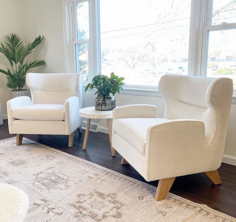 Bright living room with two cream armchairs, a small round table, large windows, and a potted plant