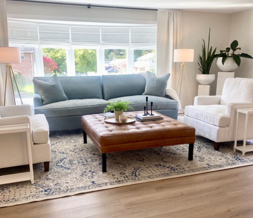 Bright living room with a gray sofa, white armchairs, wooden coffee table, and large window.