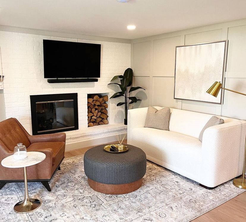 Modern living room with white sofa, brown chair, round ottoman, fireplace, TV, and neutral decor