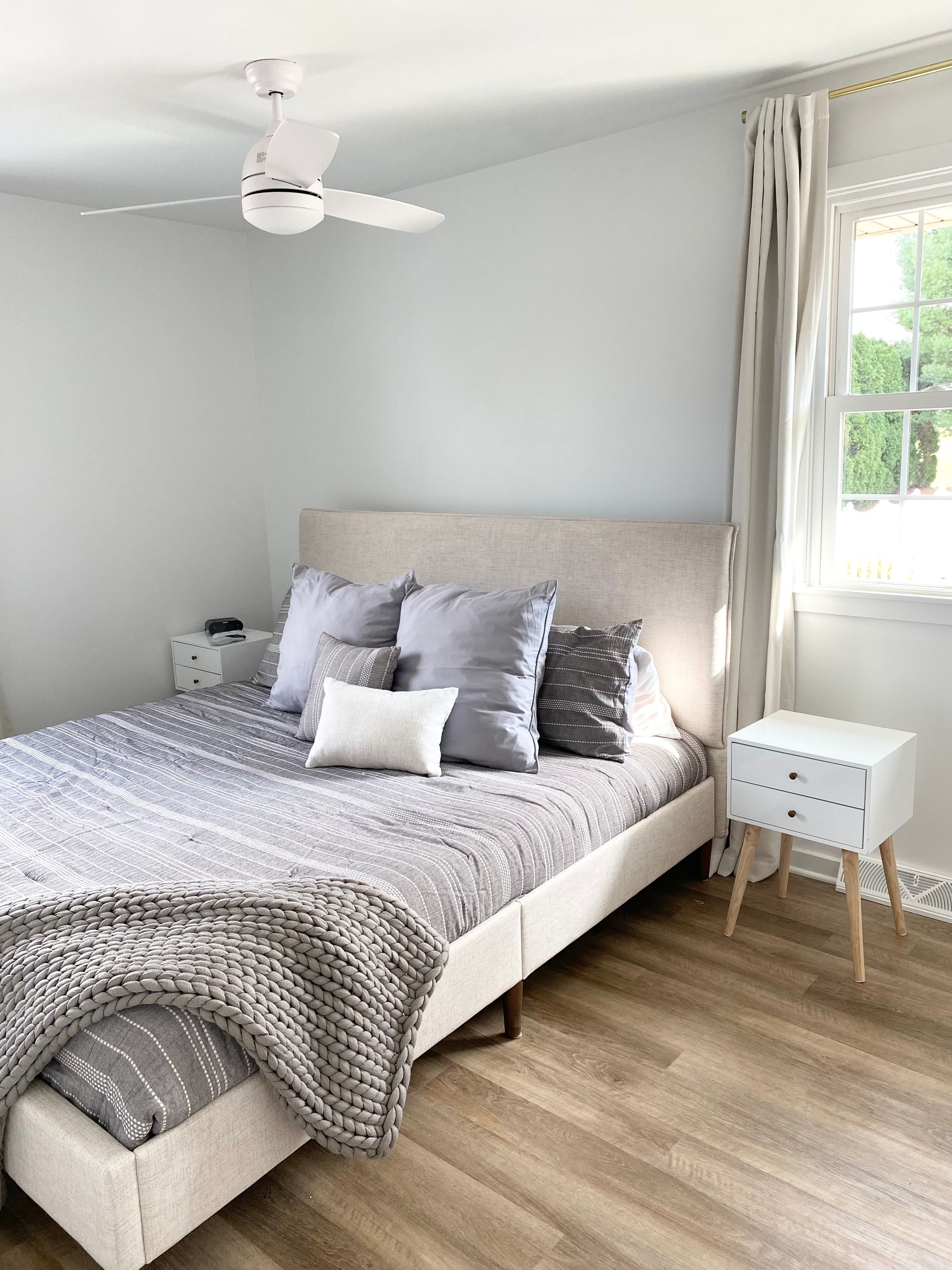 Bright bedroom with a gray bed, white nightstands, ceiling fan, and sunlight through a window.