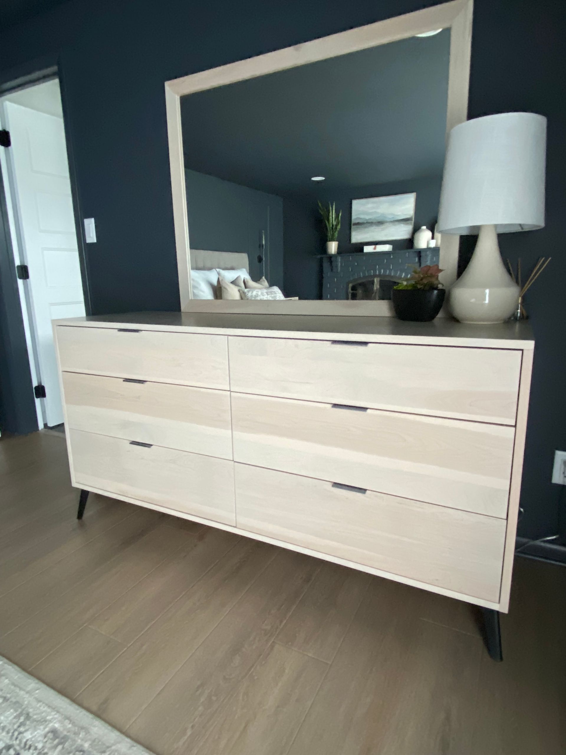 Light wood dresser with mirror in a modern bedroom, topped with a lamp and small plant.