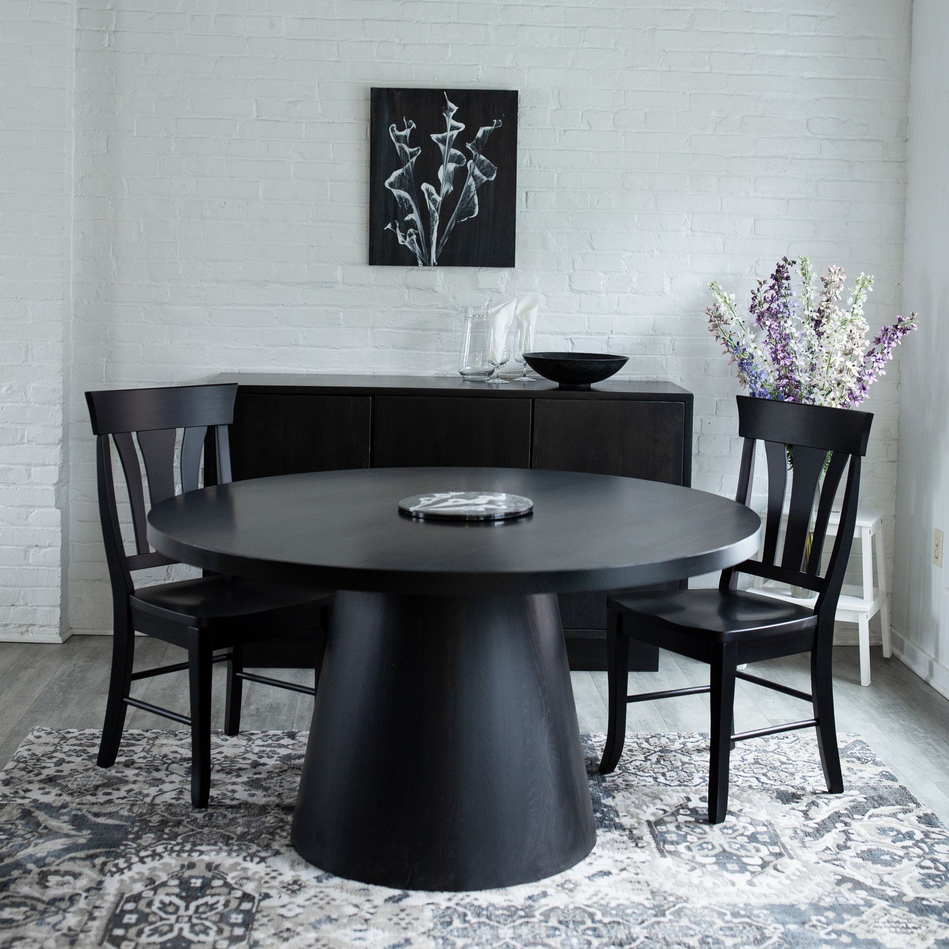 Black dining table with four chairs in a minimalist room, with wall art and a patterned rug.