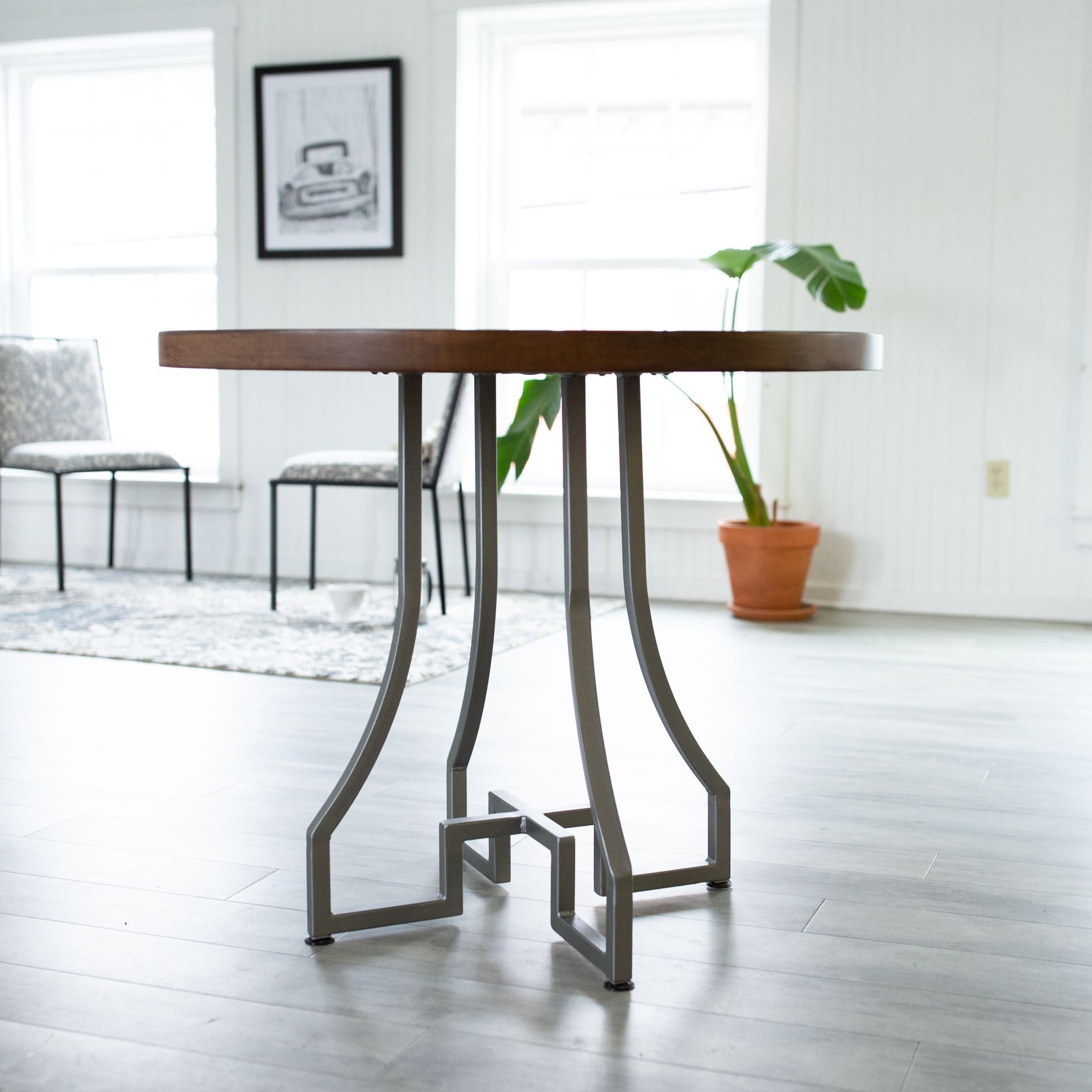Modern wooden table with metal legs in a bright room with chairs, framed art, and a potted plant.