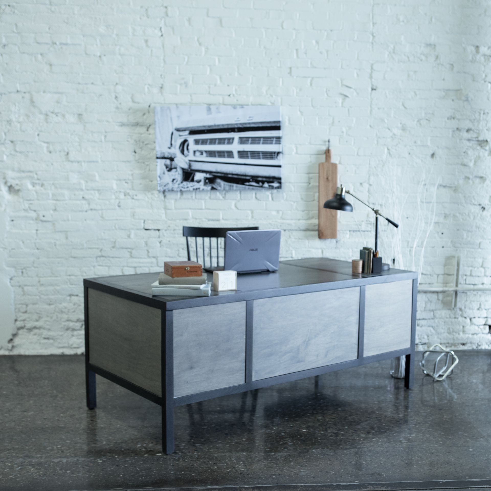 Minimal office desk in a white brick room with a laptop, lamp, books, and framed wall art