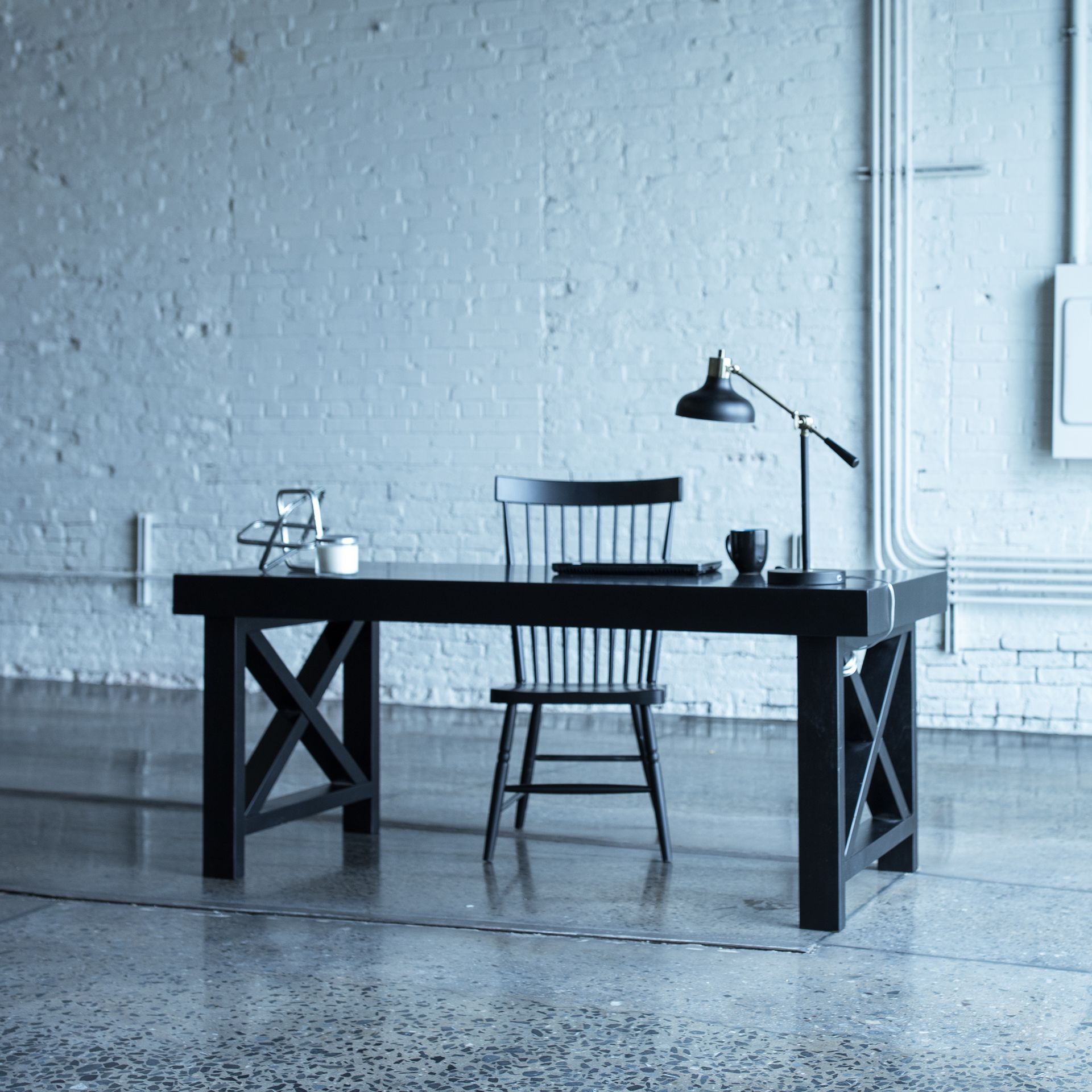 Minimalist office with a dark desk, chair, lamp, and cup on a concrete floor against a white brick wall.