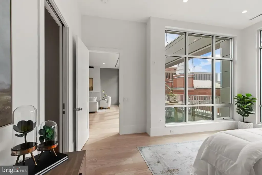 Bright modern bedroom with large windows, white bedding, and a view of neighboring houses outside.