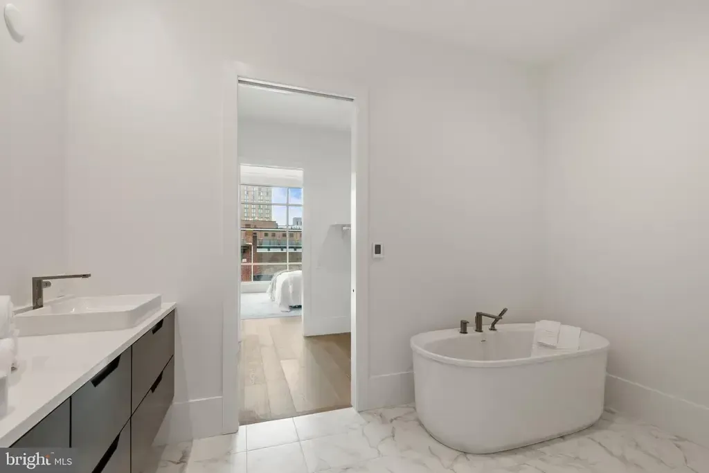 Minimalist white bathroom with vanity, freestanding tub, and doorway to a bright room beyond