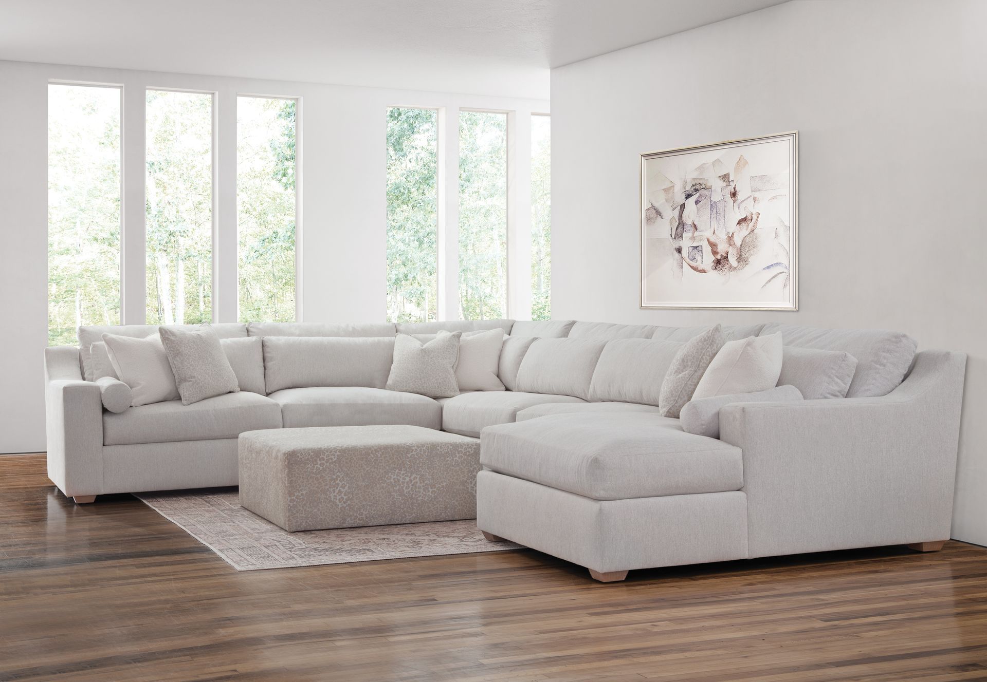 Bright living room with a large white sectional sofa, ottoman, wood floor, and tall windows.