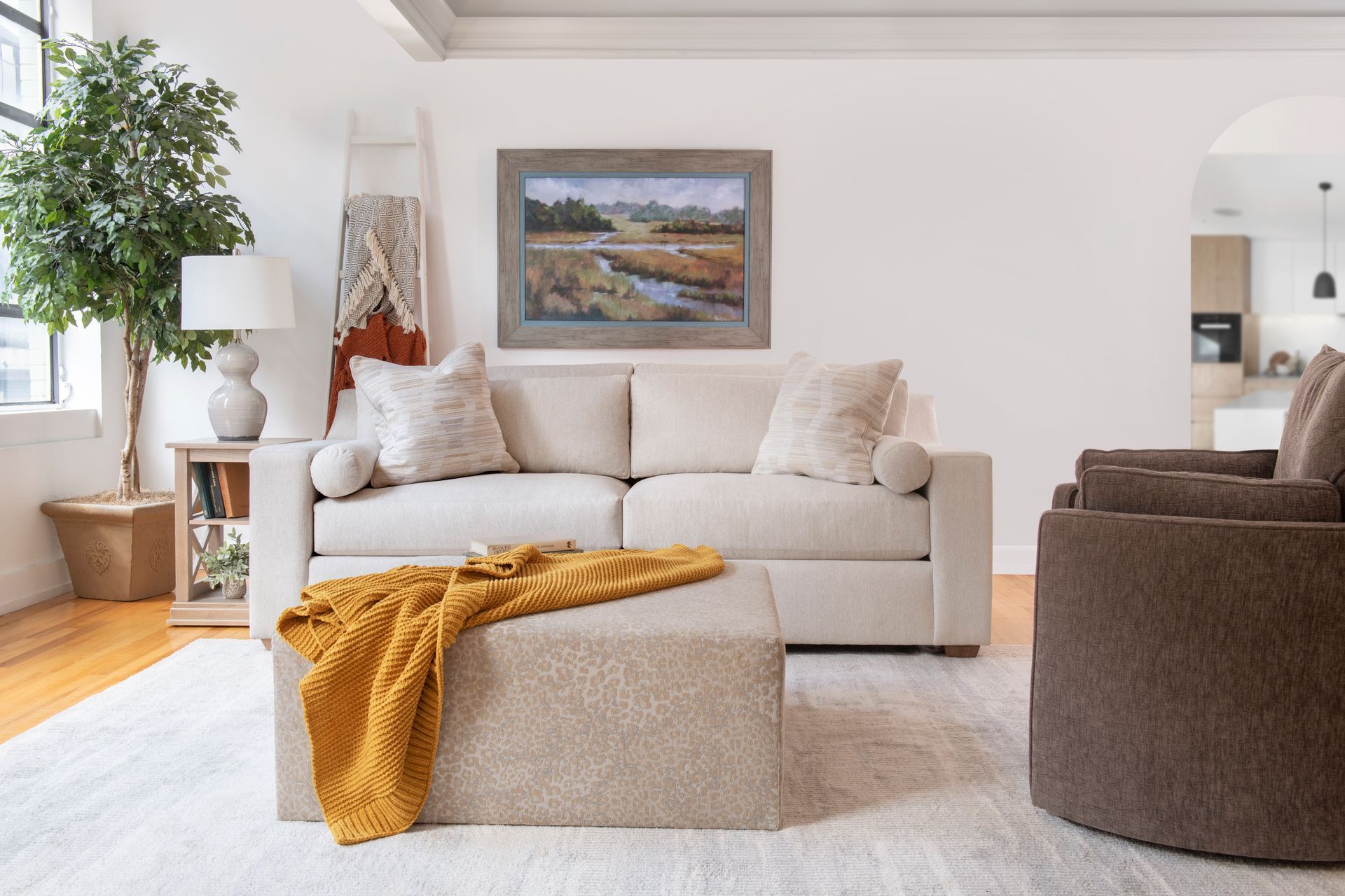 Bright living room with beige sofa, mustard throw, framed landscape, and round brown chair on a white rug