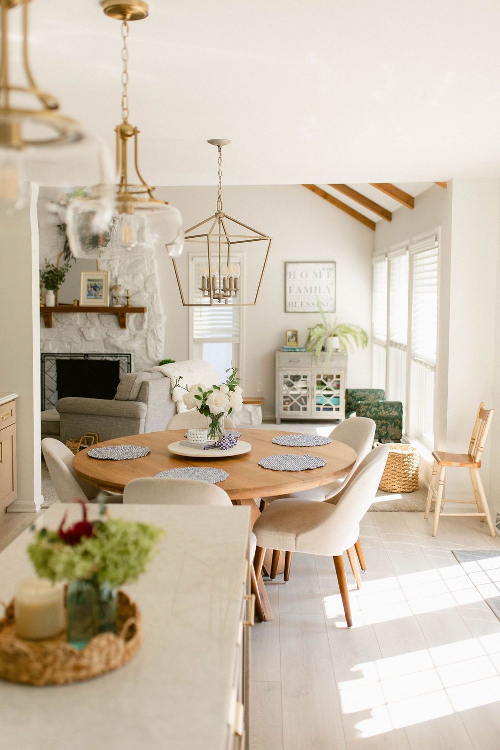 Bright, cozy dining room with a round wooden table, cream chairs, white decor, and sunlight by large windows