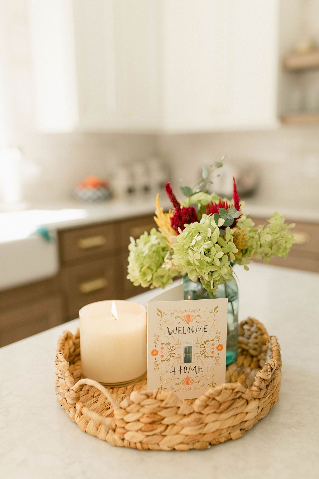 Wicker tray with white candle and floral candle jar on a kitchen counter with a blurred vase of flowers behind.