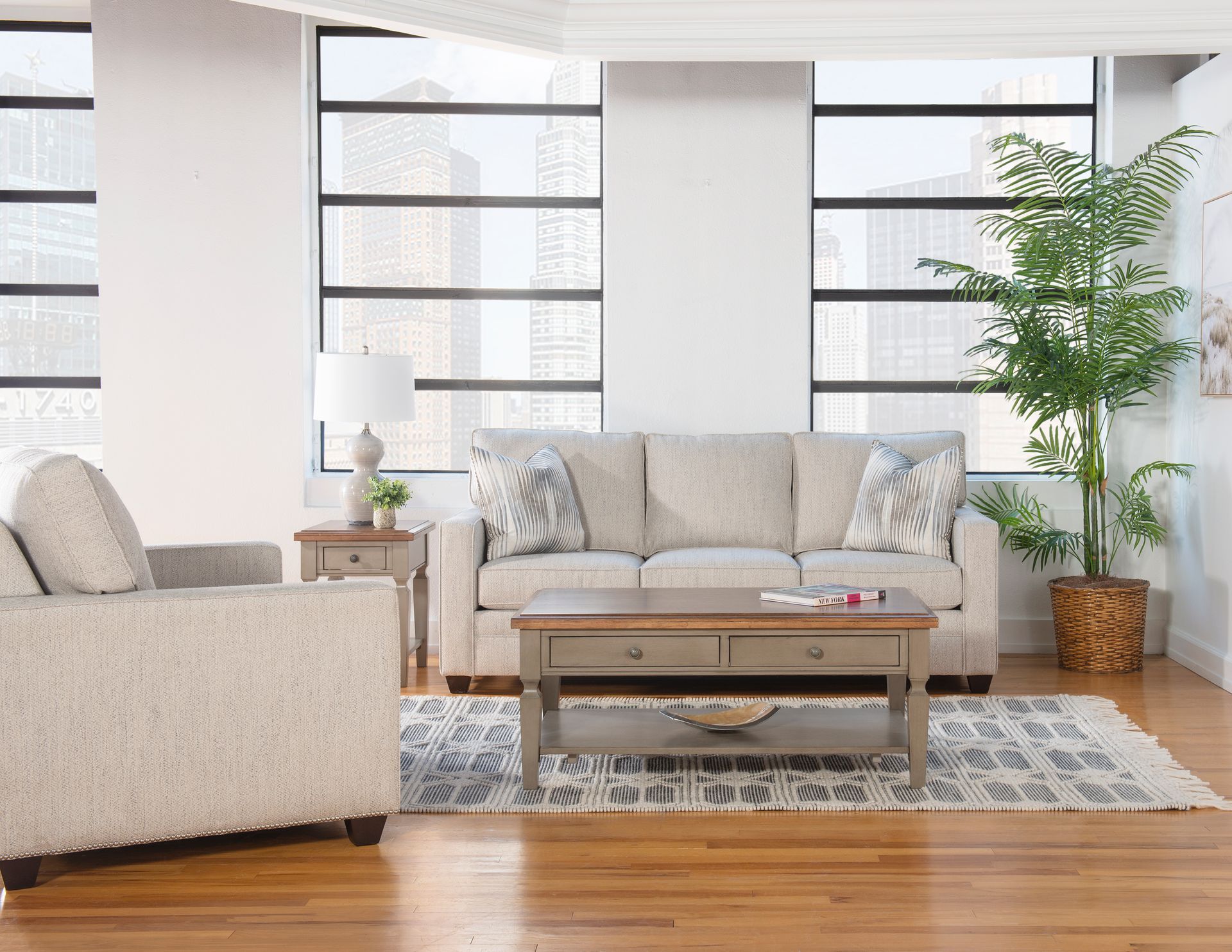 Bright living room with beige sofa, coffee table, patterned rug, and large windows with plants
