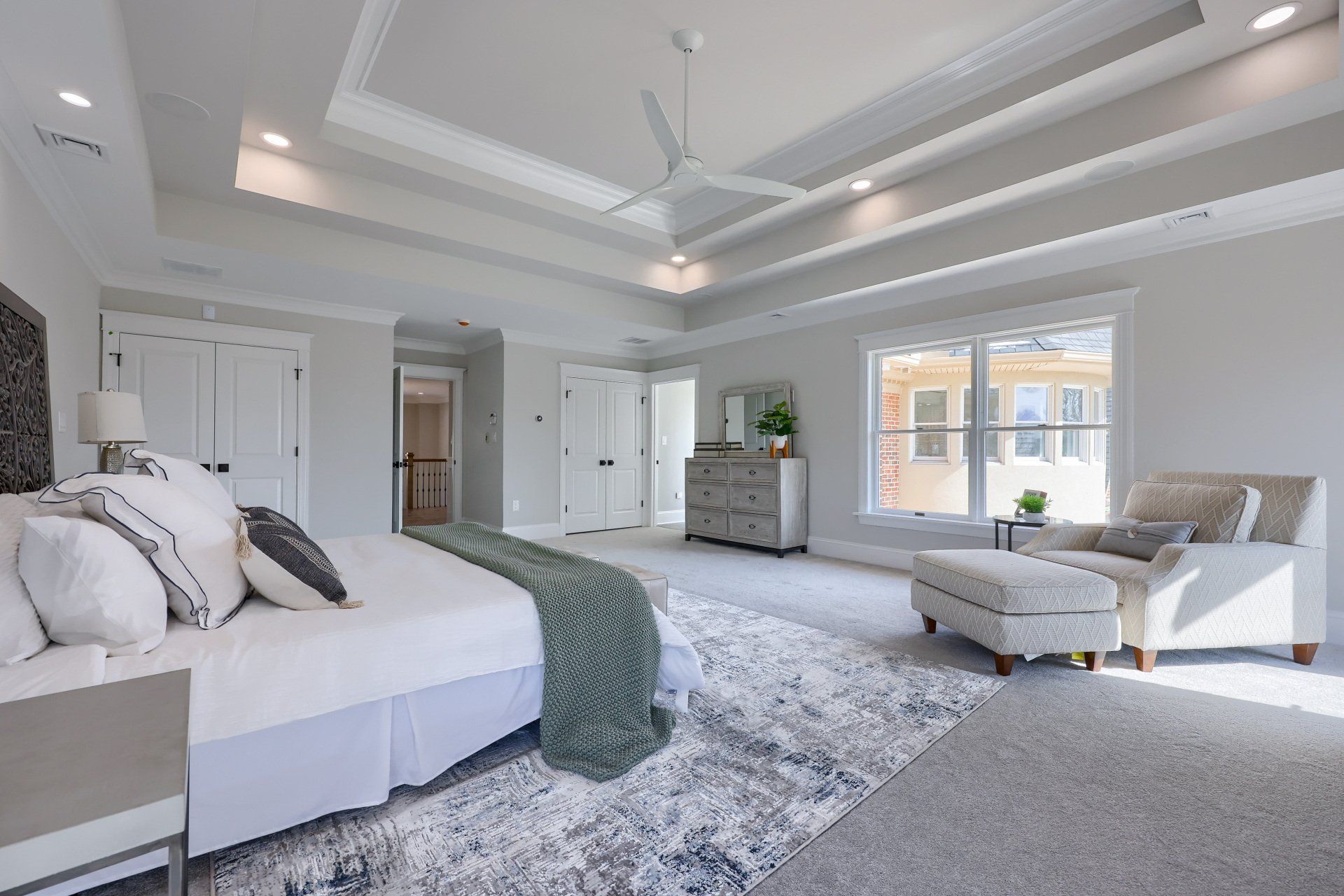 Bright modern bedroom with a king bed, gray seating, patterned rug, and large windows.