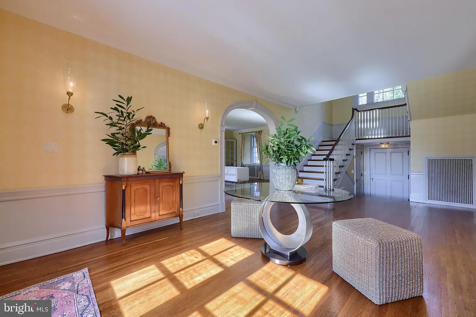 Bright foyer with hardwood floors, staircase, arched doorway, and sunlight streaming across the room.