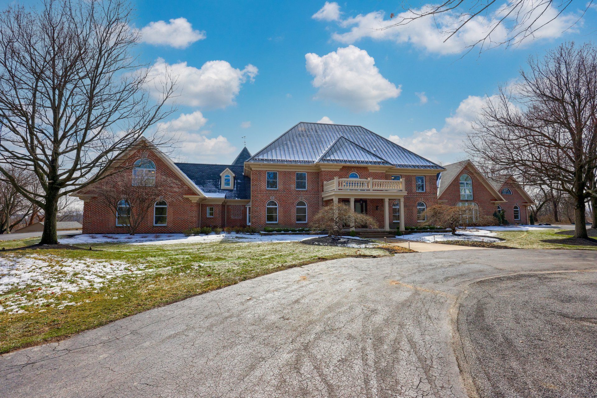 Large brick house with a snowy driveway and bare trees under a blue sky