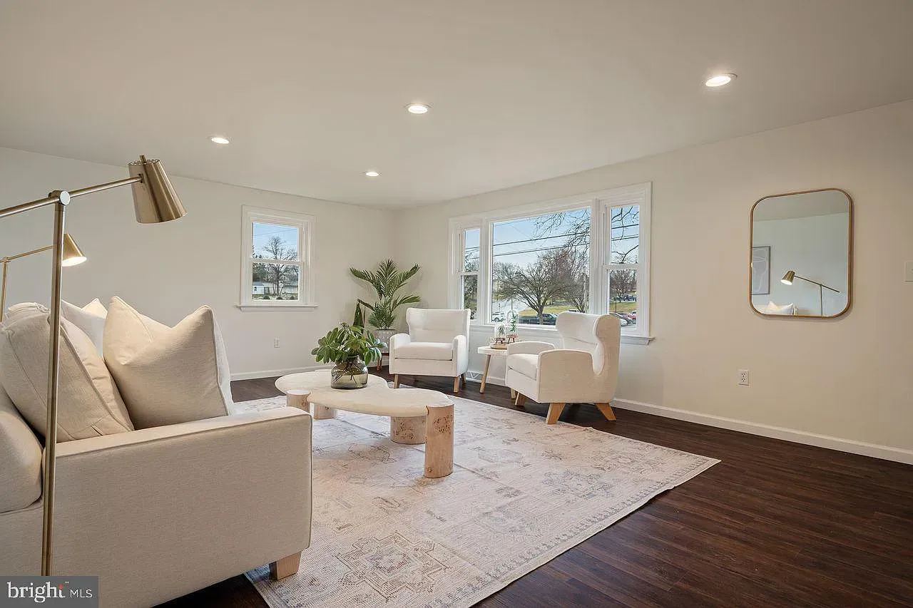 Bright living room with beige sofa, white chairs, coffee table, rug, and wall mirror on dark wood floor