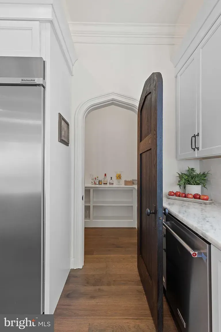 Narrow white kitchen hallway with wood floors, arched doorway, and built-in shelves at the far end