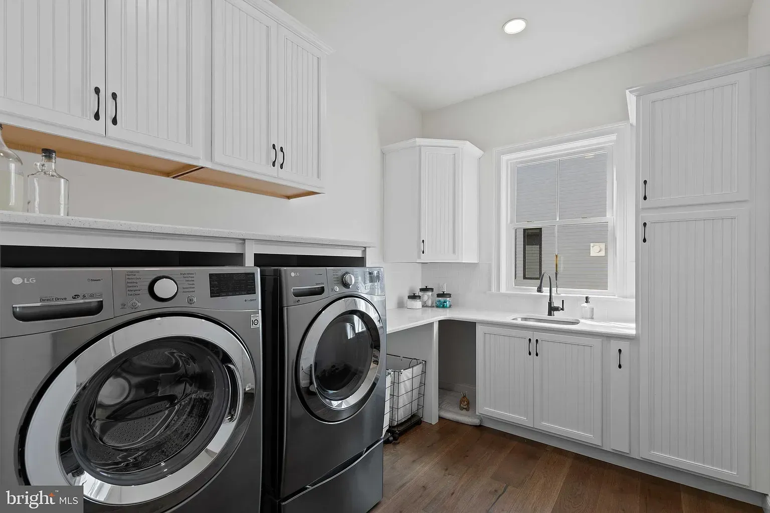 Bright laundry room with gray washer and dryer, white cabinets, sink, and wood floors