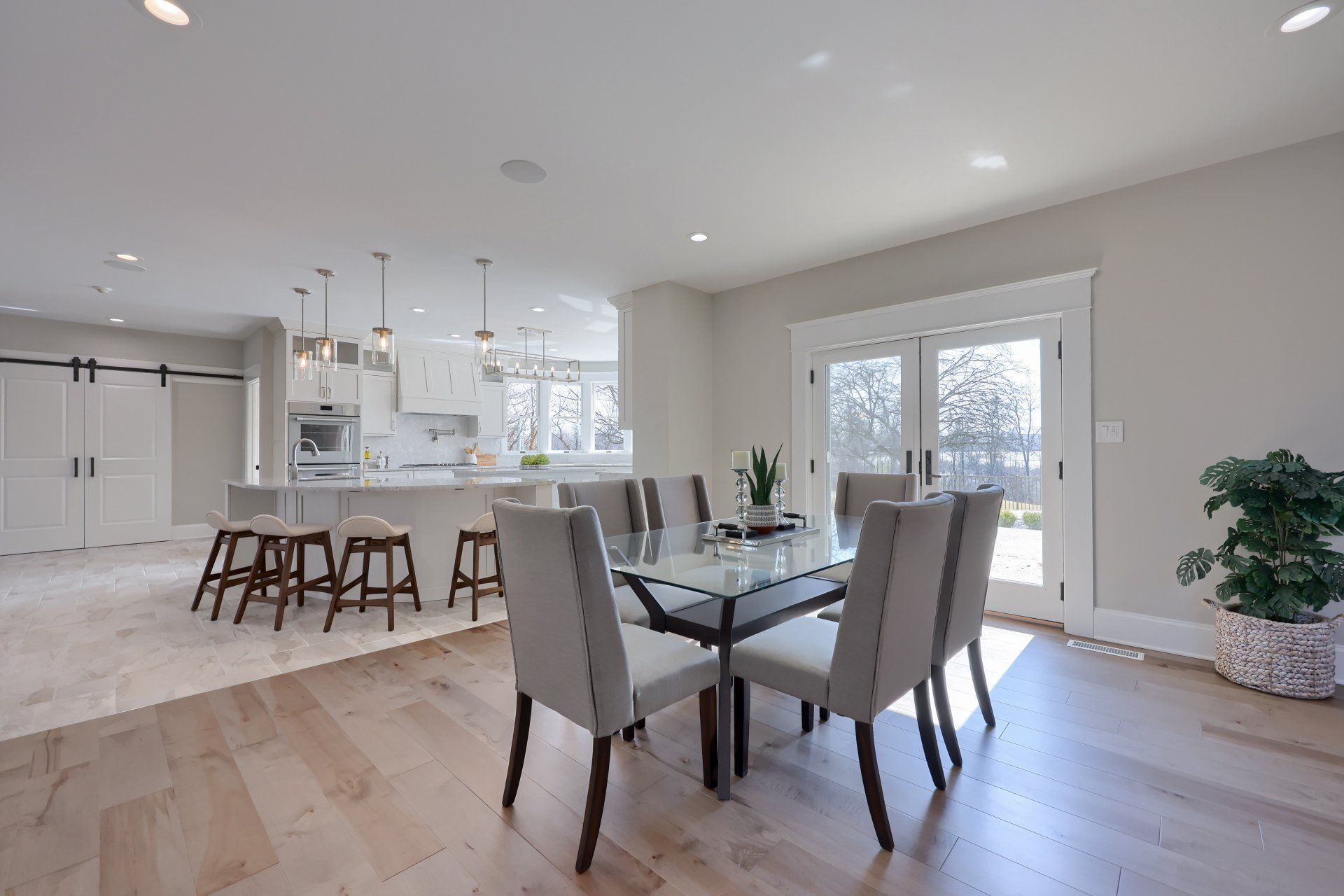 Bright modern dining area with glass table, gray chairs, kitchen barstools, and French doors to snowy outside.