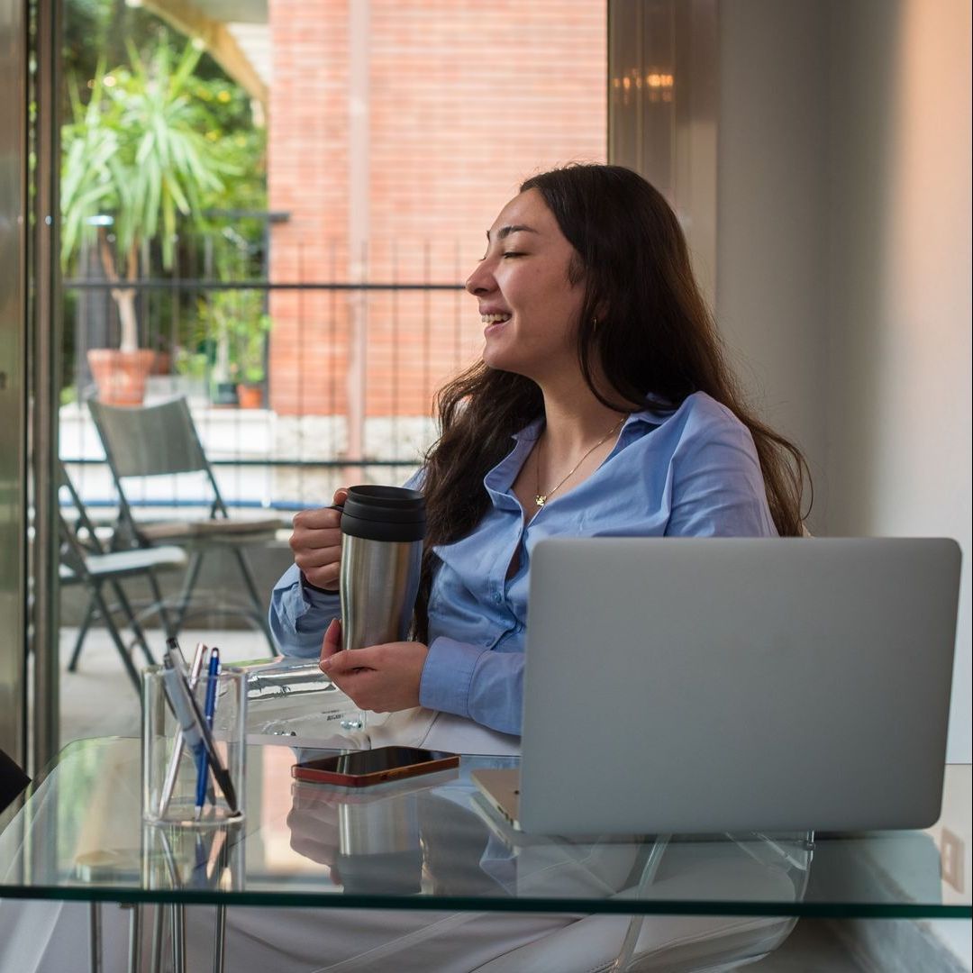 Woman holding tumbler, sitting in front of her laptop