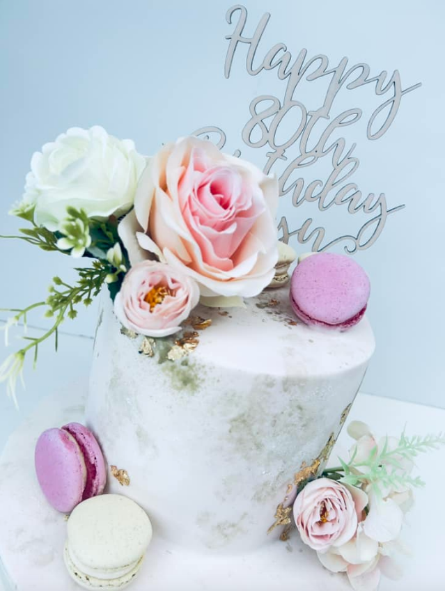 White birthday cake with pink roses, macarons, and a “Happy Birthday” topper
