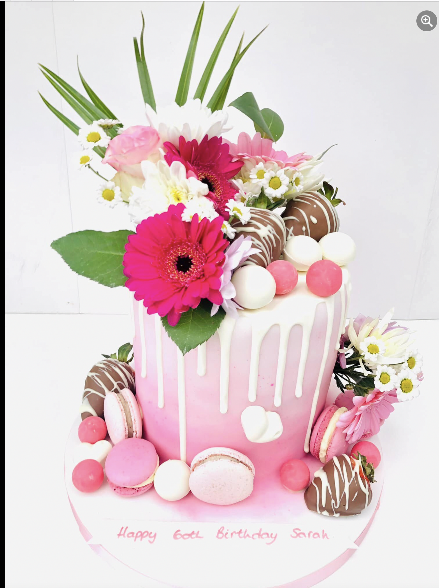 Pink birthday cake with white drip icing, bright flowers, and macarons on a white plate