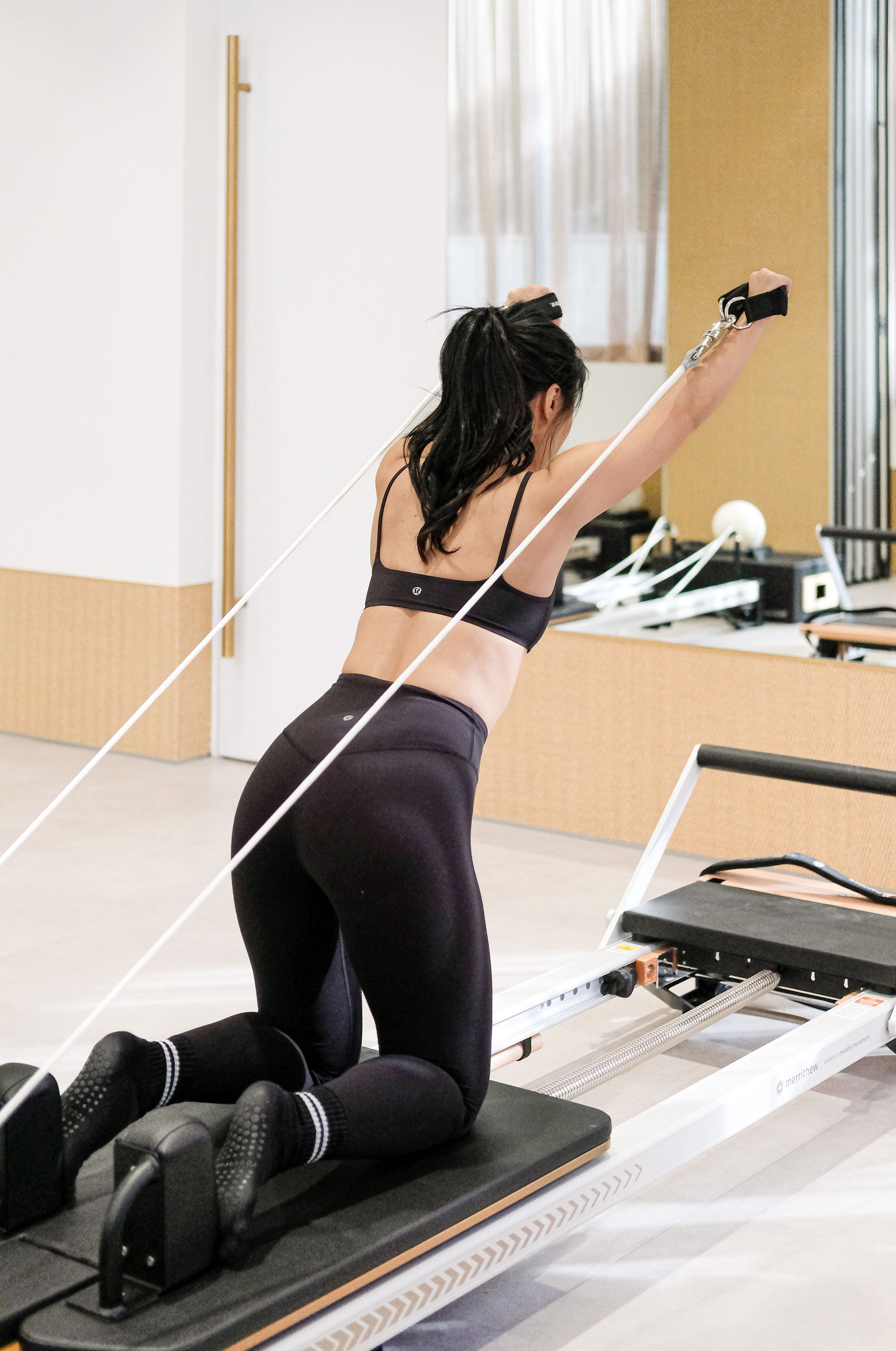 Person kneels on a reformer machine in a bright Pilates studio, reaching forward during exercise.