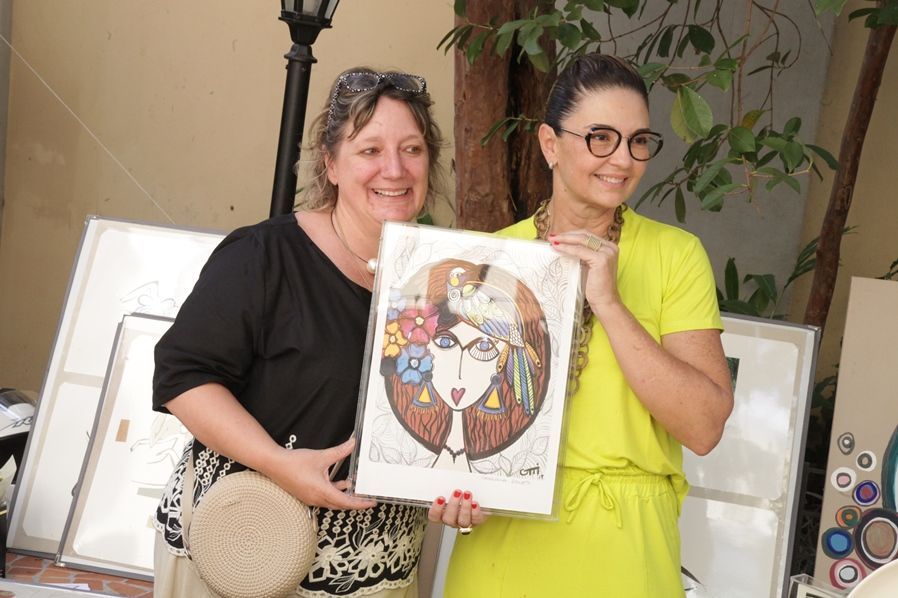 Duas mulheres estão de pé, uma ao lado da outra, segurando uma pintura do rosto de uma mulher.