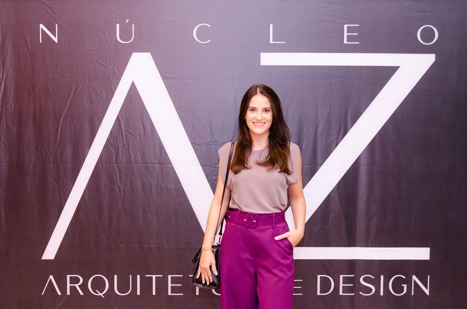 Uma mulher de calça roxa está parada em frente a uma placa que diz nucleo arquitetura design.