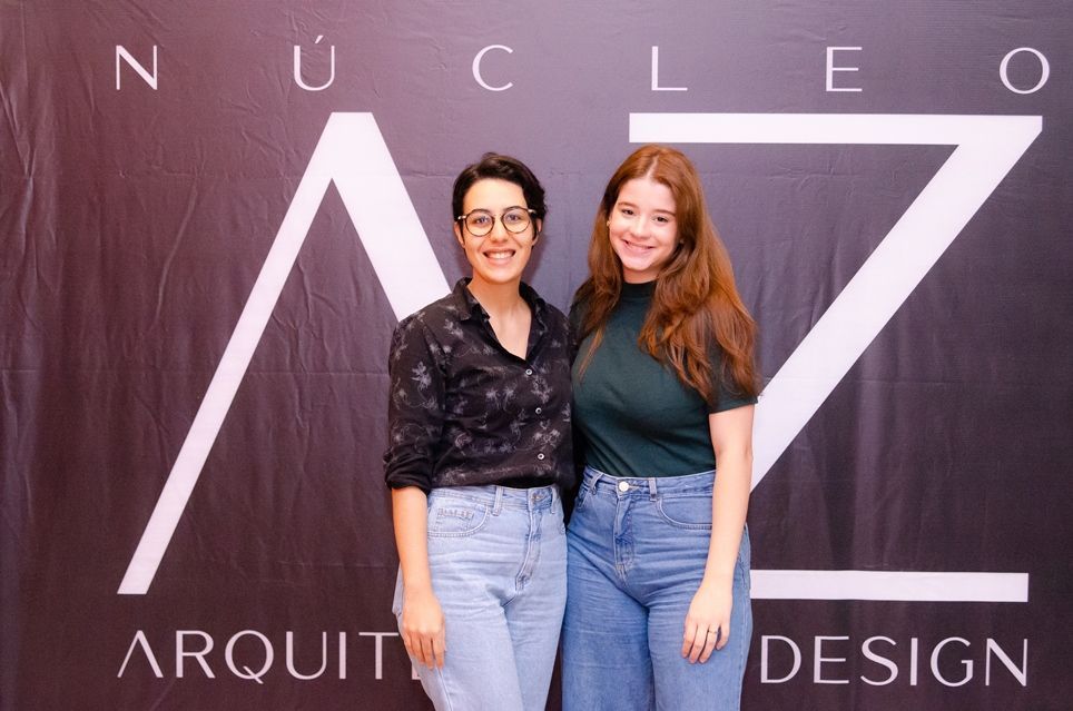 Duas mulheres posam para uma foto em frente a uma placa que diz nucleo arquit design