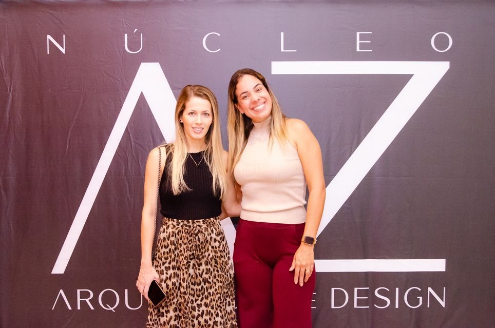 Duas mulheres posam para uma foto em frente a uma placa que diz nucleo arqui design